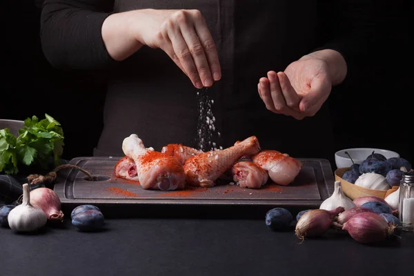 Una chef espolvorea palitos de pollo crudos frescos sobre un fondo oscuro con sal marina. Cerca se encuentran los ingredientes para cocinar: chalotas, ciruelas azules, ajos, sal y perejil — Foto de Stock