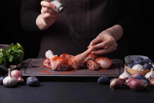 Una chef espolvorea palitos de pollo crudos frescos sobre un fondo oscuro con sal marina. Cerca se encuentran los ingredientes para cocinar: chalotes, ciruelas azules, ajos, s — Foto de Stock