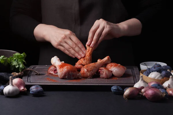 Mujer chef frota la sal con los palillos de pollo crudo fresco sobre un fondo oscuro. Cerca se encuentran los ingredientes para cocinar: chalotas, ciruelas azules, ajos, sal y perejil — Foto de Stock