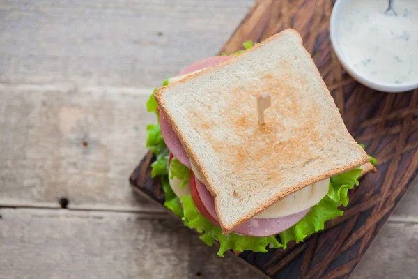 Close-up big sandwich with ham, cheese, tomatoes, salad and white sauce on toasted bread on a rustic wooden table. Top view with copy space — Stock Photo, Image