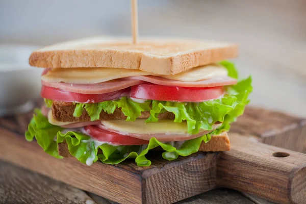 Yakın çekim büyük sandviç jambon, peynir, domates, salata ve rustik ahşap bir masa üzerinde kızarmış ekmek üzerinde beyaz sos ile