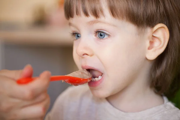 Bellissimo bambino mangia porridge dalla mano della mamma. La madre nutre il figlio con un cucchiaio. Ragazzo con lunghi capelli biondi e grandi occhi blu probabilmente — Foto Stock