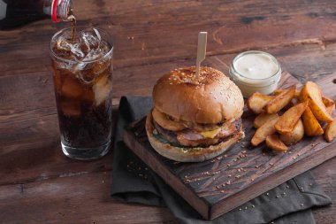 Aperatifler beyaz Sarımsak soslu patates şeklinde ve soğuk kola bir bardak ile lezzetli bir Burger. soğan halkaları, pastırma ve koyu ahşap arka plan üzerinde peynir ile sulu Burger