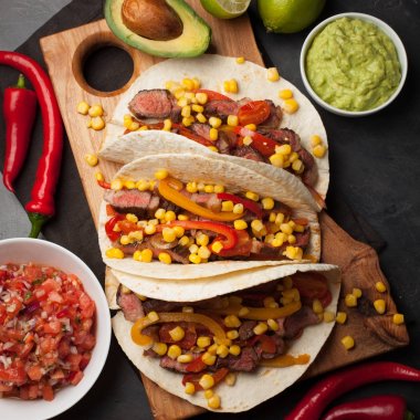 Mermer sığır eti, siyah Angus ve karanlık bir taş zemin üzerine ahşap tahta üzerinde sebze ile üç Meksikalı Taco. Soslar guacamole ve kase salsa Meksika tabak. Üstten Görünüm