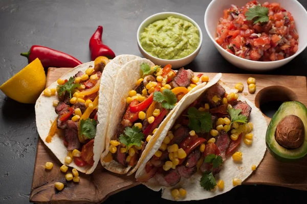 Mermer sığır eti, siyah Angus ve karanlık bir taş zemin üzerine ahşap tahta üzerinde sebze ile üç Meksikalı Taco. Soslar guacamole ve salsa kase ile Meksika yemek