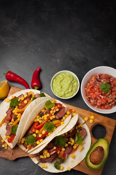 Tre mexikanska tacos med marmorerat nötkött, black Angus och grönsaker på träskiva på en mörk sten bakgrund. Mexikansk maträtt med såser guacamole och salsa i skålar. Ovanifrån med kopia utrymme — Stockfoto