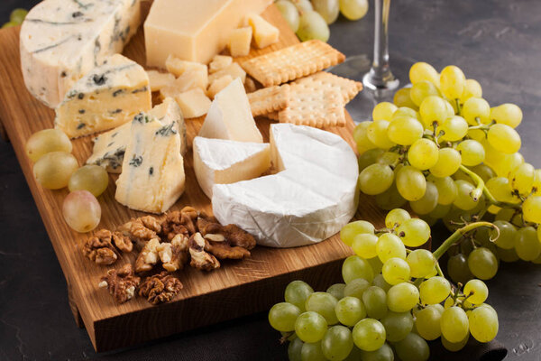 Assorted cheeses with white grapes, walnuts, crackers and white wine on a wooden Board. Food for a romantic date on a dark stone background. Top view