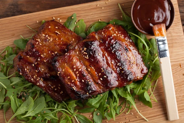 Closeup de costelas de porco grelhadas com molho de churrasco e caramelizadas em mel em uma cama de arugula. Lanche saboroso à cerveja em uma tábua de madeira da submissão no velho contexto de madeira. Vista superior — Fotografia de Stock