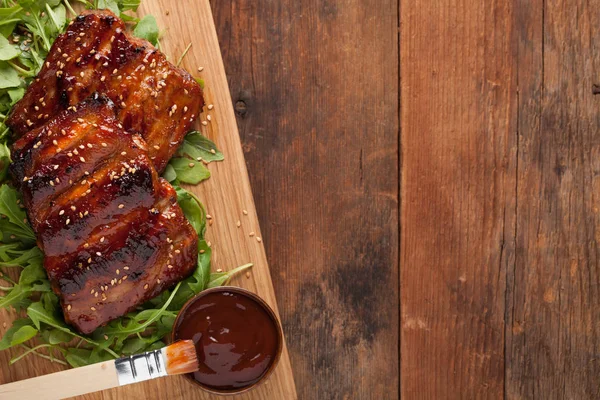 Primo piano di costolette di maiale grigliate con salsa barbecue e caramellate nel miele su un letto di rucola. Spuntino saporito a birra su un'asse di legno per sottomissione sullo sfondo vecchio di legno. Vista dall'alto con spazio di copia — Foto Stock