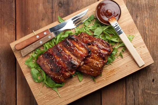 Closeup de costelas de porco grelhadas com molho de churrasco e caramelizadas em mel em uma cama de arugula. Lanche saboroso à cerveja em uma tábua de madeira da submissão no velho contexto de madeira. Vista superior — Fotografia de Stock