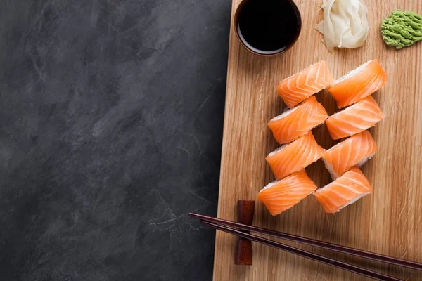 Ένα κλασικό Φιλαδέλφεια ρολό με wasabi, τζίντζερ και σάλτσα σόγιας για μια ξύλινη σανίδα. Σολομός, τυρί Φιλαδέλφεια, αβοκάντο, αγγούρι. Ιαπωνική Σούσι σε σκούρο φόντο πέτρα. Το Top view με αντίγραφο χώρου — Φωτογραφία Αρχείου