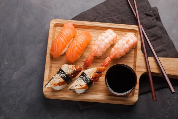 Un set di sushi con salmone, gamberetti e anguilla con una ciotola di salsa di soia e bacchette di legno su una tavola di legno. Delizioso cibo giapponese su uno sfondo di pietra scura. Vista dall'alto — Foto Stock