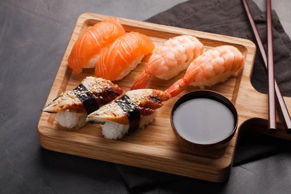 Un set di sushi con salmone, gamberetti e anguilla con una ciotola di salsa di soia e bacchette di legno su una tavola di legno. Delizioso cibo giapponese su uno sfondo di pietra scura. Vista dall'alto — Foto Stock