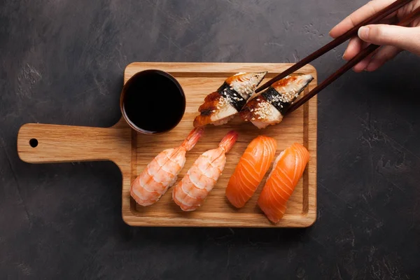 Un juego de sushi con salmón, camarones y anguila y palillos de madera en una tabla de madera. Deliciosa comida japonesa sobre un fondo de piedra oscura. La mano femenina toma los palillos en el sushi. Vista superior — Foto de Stock