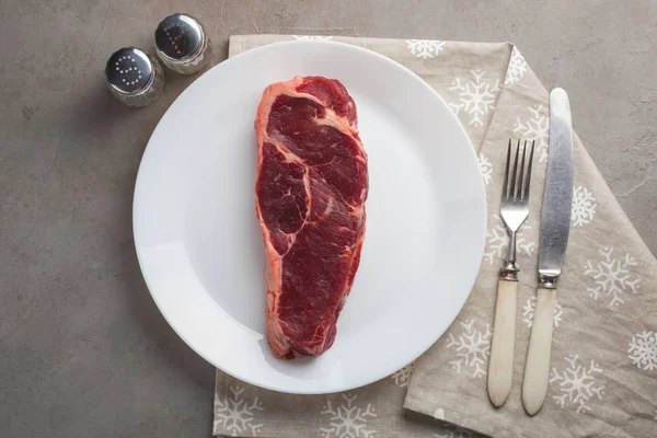 Syrové hovězí maso mramorované steak s vintage bílé nádobí na staré kamenné pozadí. Kousek masa s pepřem a solí na stole. Steak z New Yorku. Pohled shora — Stock fotografie