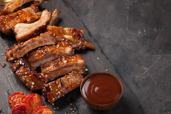 Costolette di maiale in salsa barbecue e pomodori arrosto al miele su un piatto di ardesia nera. Un ottimo snack per la birra su uno sfondo di pietra nera. Vista dall'alto con spazio di copia — Foto Stock