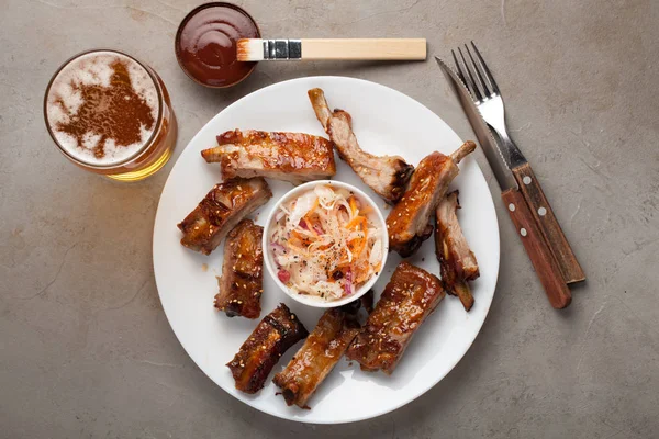 Grilled pork ribs in barbecue sauce and honey with sauerkraut and beer on a white plate. Snack to beer on a light stone table. Top view — Stock Photo, Image