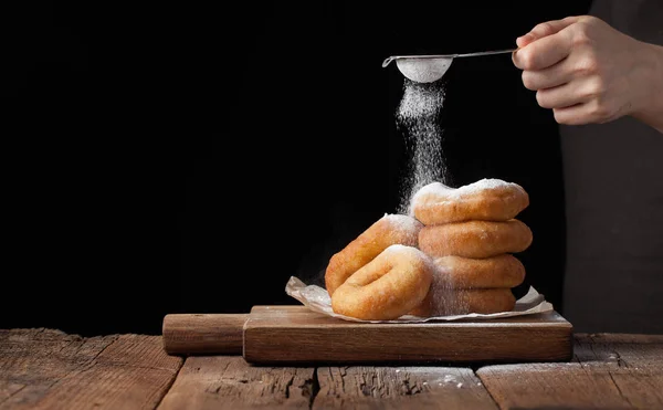 Пекарь посыпать сладкие пончики с сахаром порошка на черном фоне. Вкусная, но вредная еда на старом деревянном столе с копировальным пространством — стоковое фото