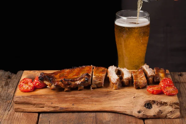 Pork ribs in barbecue sauce and honey baked tomatoes on the old wooden table. Meats and light beer on black background with copy space. In the background poured the beer into the glass — Stock Photo, Image