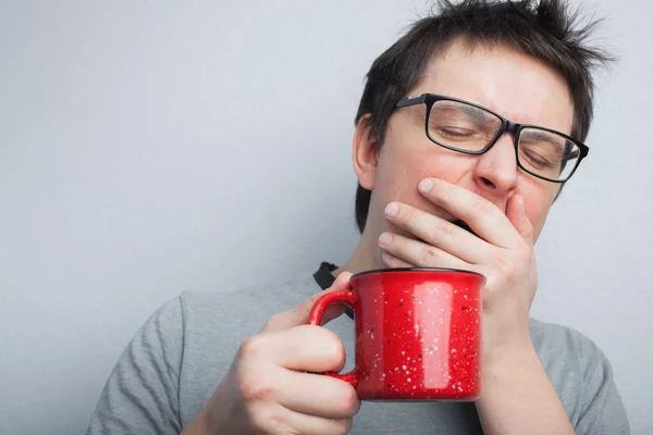 El hombre bostezo soñoliento en gafas con taza roja de té o café tiene el pelo sin peinar en ropa interior sobre fondo claro, refresco matutino y bebida. Copia espacio para tu texto — Foto de Stock