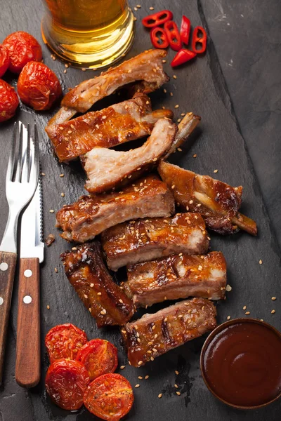 Costeletas de porco em molho de churrasco e mel tomates assados e um copo de cerveja em um prato de ardósia preta. Um ótimo lanche para cerveja em um fundo de pedra escura. Vista superior — Fotografia de Stock