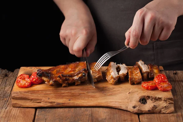 O chef corta com uma faca afiada pronta para comer costelas de porco, deitada em uma velha mesa de madeira. Um homem prepara um lanche para cerveja em um fundo preto com espaço de cópia — Fotografia de Stock