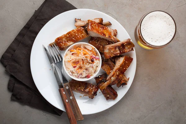 Grilled pork ribs in barbecue sauce and honey with sauerkraut and beer on a white plate. Snack to beer on a light stone table. Top view — Stock Photo, Image