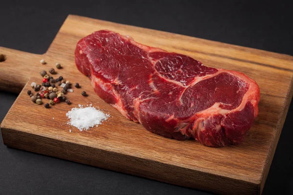 Bife de vaca no fundo preto de madeira. Fotografia horizontal. Fundo alimentar. Mármore a carne com pimenta e sal em um fundo escuro — Fotografia de Stock