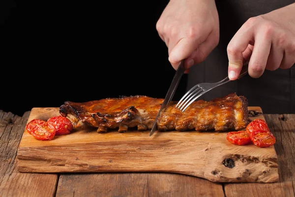 The chef cuts it with a sharp knife ready to eat pork ribs, lying on an old wooden table. A man prepares a snack to beer on a black background with copy space — Stock Photo, Image