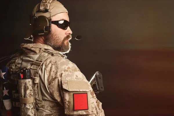 Brutal man in the military desert uniform and body armor is pathetic and looks away at the black background in the Studio. The bearded player in the airsoft safety goggles is holding his rifle