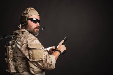 Acımasız adam askeri çöl üniforma ve vücut zırh standları ve stüdyoda siyah bir zemin üzerine silah tutar. Airsoft koruyucu gözlük ya da gözlük ve etkin kulaklık sakallı oyuncu