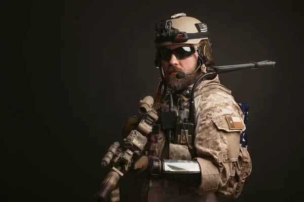 Acımasız adam askeri çöl üniforma ve vücut zırh mücadele bir rafa anlamına gelir ve bir siyah arka plan üzerine tüfeğini tutar. Amaçlayan bir tüfek airsoft koruyucu gözlük sakallı Player'da — Stok fotoğraf