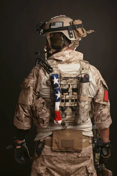 Brutale man in de militaire woestijn uniform en lichaam armor is de moeite waard zijn rug en houdt van zijn geweer op een zwarte achtergrond. De bebaarde speler in de airsoft veiligheidsbrillen — Stockfoto