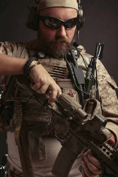 Brutal man in the military desert uniform and body armor stands and holds his rifle on a black background. The bearded player in the airsoft reloads his carabine
