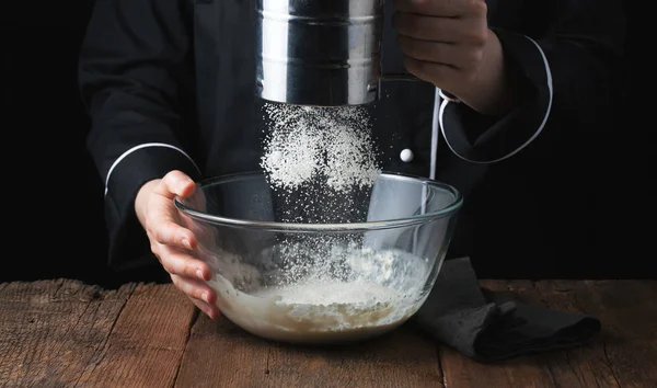 Chef manos verter harina en polvo sobre la masa cruda utilizando tamiz sobre un fondo negro, proceso de cocción — Foto de Stock