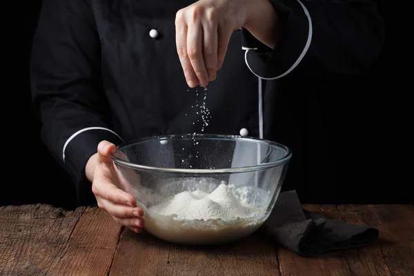 Chef manos espolvorea sal marina sobre la masa cruda sobre un fondo negro, proceso de cocción — Foto de Stock