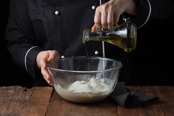 Las manos del chef vierten aceite de oliva en un tazón sobre la masa cruda sobre un fondo negro, proceso de cocción de la masa para pizza — Foto de Stock