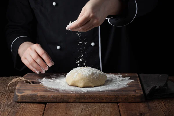 Chef manos verter harina en polvo sobre la masa cruda utilizando tamiz sobre un fondo negro, proceso de cocción — Foto de Stock