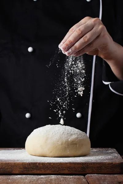 Chef manos verter harina en polvo sobre la masa cruda utilizando tamiz sobre un fondo negro, proceso de cocción — Foto de Stock