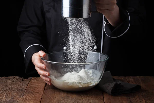 Chef manos verter harina en polvo sobre la masa cruda utilizando tamiz sobre un fondo negro, proceso de cocción — Foto de Stock