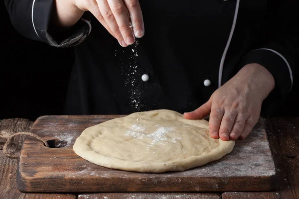 Руки шеф-повара наливают порошок муки на сырое тесто с помощью сита на черном фоне, процесс приготовления — стоковое фото