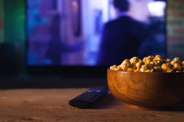 Una ciotola di popcorn in legno e telecomando sullo sfondo funziona la TV. Sera accogliente guardare un film o serie TV a casa — Foto Stock