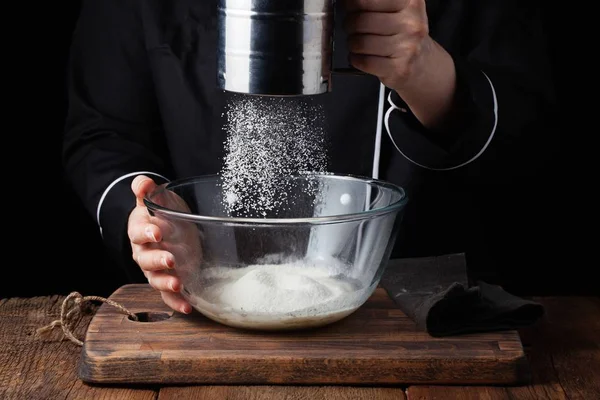 Chef manos verter harina en polvo sobre la masa cruda utilizando tamiz sobre un fondo negro, proceso de cocción — Foto de Stock