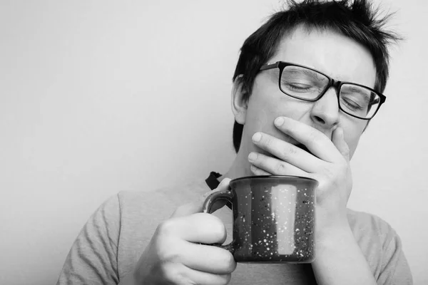 El hombre bostezo soñoliento en gafas con taza roja de té o café tiene el pelo sin peinar en ropa interior sobre fondo claro, refresco matutino y bebida. Copia espacio para tu texto. foto en blanco y negro — Foto de Stock