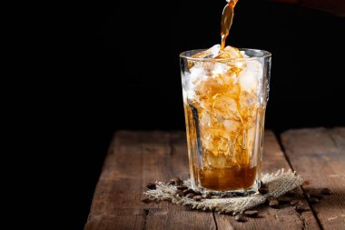 Buzlu kahve üzerinden uzun bir cam ve kahve çekirdekleri bir eski rustik ahşap tablo. Kopya alanı ile karanlık bir arka plan üzerinde soğuk yaz içeceği. Bir bardak içine bir kahve kabı üzerinden dökülen içki süreci