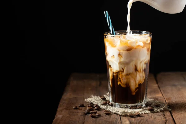 Es kopi dalam gelas tinggi dengan krim dituangkan di atas dan biji kopi di atas meja kayu pedesaan tua. Minuman musim panas yang dingin di latar belakang kayu gelap dengan ruang fotokopi — Stok Foto