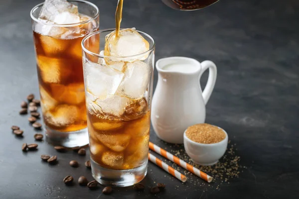 Es kopi dalam gelas tinggi di atas dan biji kopi di atas meja kayu pedesaan tua. Minuman musim panas yang dingin dengan latar belakang gelap dengan ruang fotokopi. Proses menuangkan minuman dari teko kopi ke dalam gelas — Stok Foto