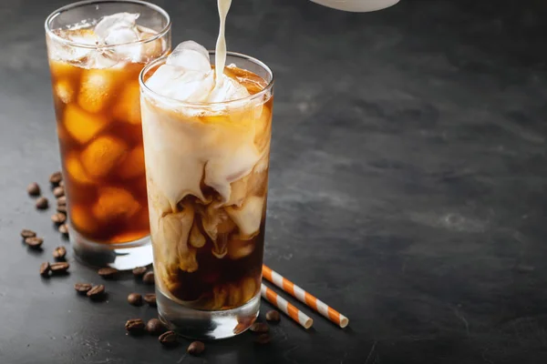 Ice coffee in a tall glass with cream poured over, brown sugar and coffee beans. Cold summer drink on a dark background. With copy space