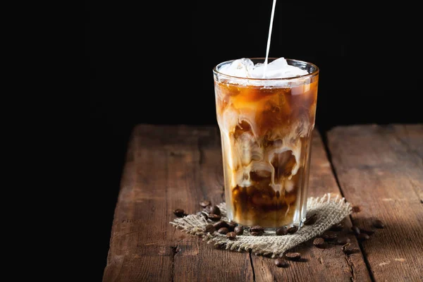 Es kopi dalam gelas tinggi dengan krim dituangkan di atas dan biji kopi di atas meja kayu pedesaan tua. Minuman musim panas yang dingin di latar belakang kayu gelap dengan ruang fotokopi — Stok Foto