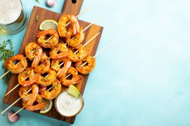 Açık mavi beton zemin üzerinde limon, sarımsak ve sosla servis edilen ızgara karides şişleri veya langoustinler. Deniz ürünleri ve bira. Kopyalama alanı olan üst görünüm. Düz yatıyordu